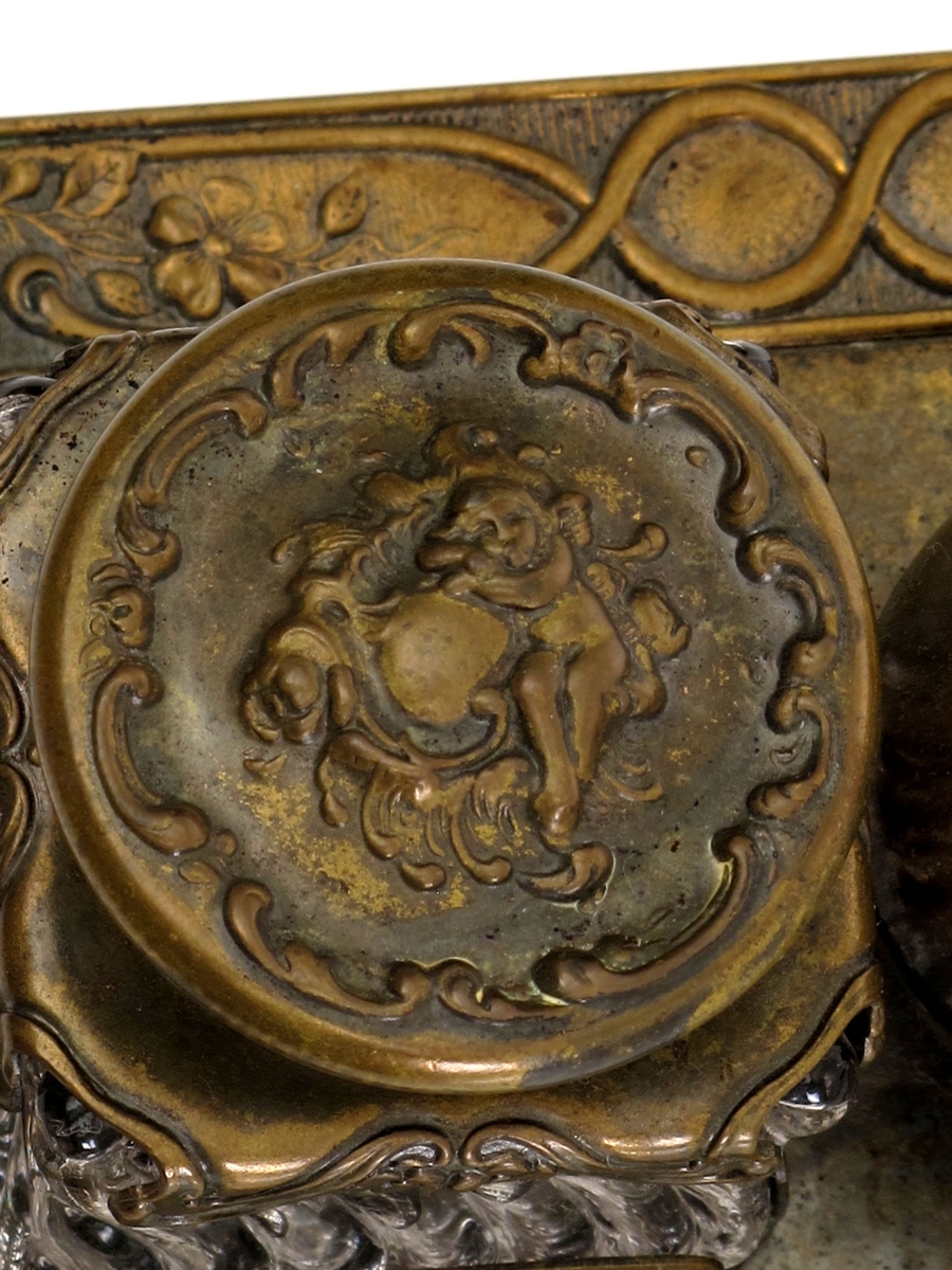 Skrivestell. Presset  messing, 2 blekkhus av  pressglass. Rektangulær messingplate med presset ornamentbord langs kanten og 4 små dreide føtter. Presset dekorativt mønster.