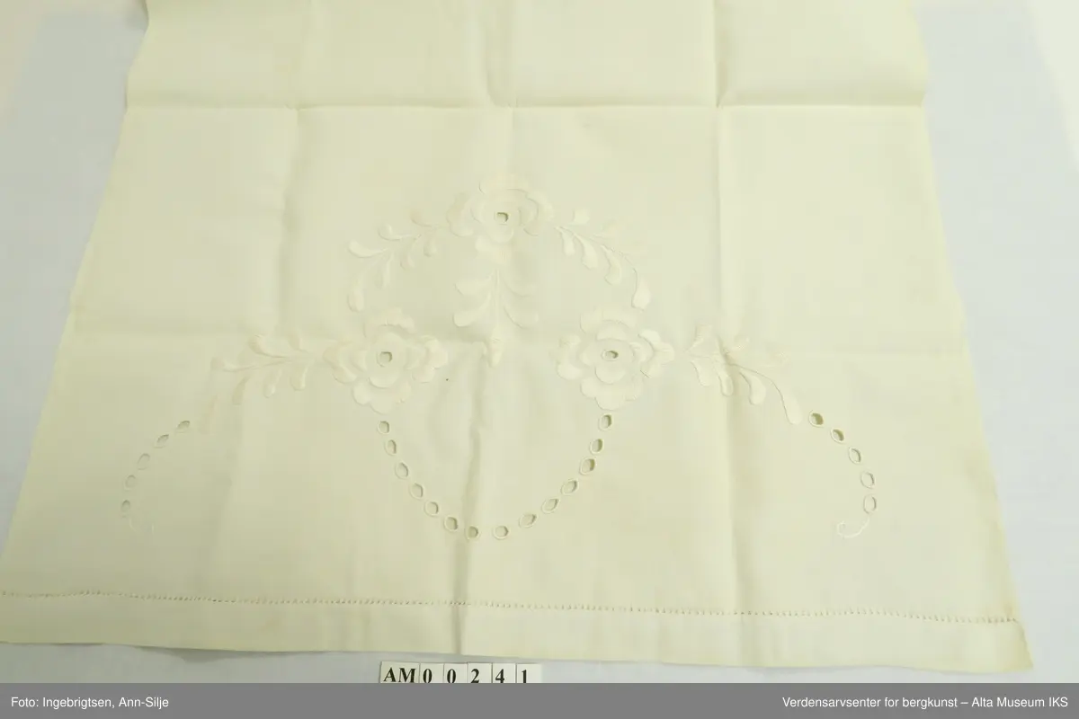 Stort pyntehåndkle med broderi i hver ende. Det ene broderiet er større enn det andre. Det store broderiet er av blomster og blader, mens det minste ser ut til å være et monogram eller lignende.