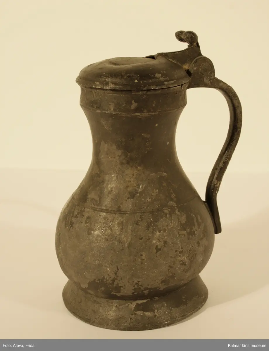 KLM 1886. Kanna, hankkärl. Av tenn. Hankkärl, bukig form, försedd med lock och handtag men utan näbb. Grepen ornerad på utsidan med bladranka som upptill avslutas med en djurliknande figur. Bottenmedalj i kannan, i form av korsfästelsemedalj. Spår efter förlorad lockmedalj, på lockets insida.