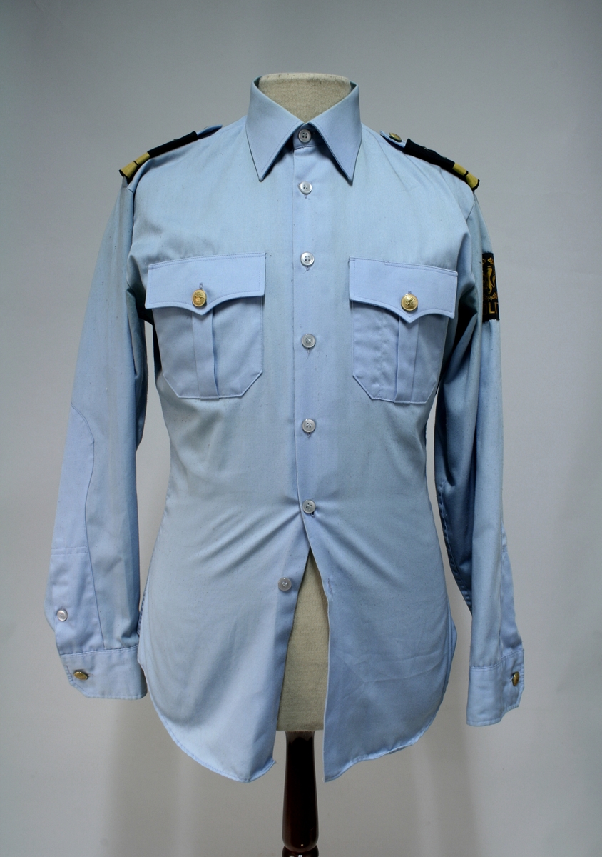 Uniform for politinspektør etter 1963-reglement med merking etter 1977-reglement. Helårsuniform M/1963 med skjorte, langbukse, belte, helårskappe og jakke. Skyggelue med gullbrodert bånd, loslue og båtlue M/1967. Kappen og jakken har skulderklaffer av flettede gullsnorer med to sølvstjerner og festeknapp i gullbroderi. Skjorten har en enklere skulderklaff med to gule render og to sølvstjerner. Det følger med et ekstra enkelt lærbelte og et ekstra sett skulderklaffer til skjorte. Plaggene har gullfargede løveknapper.