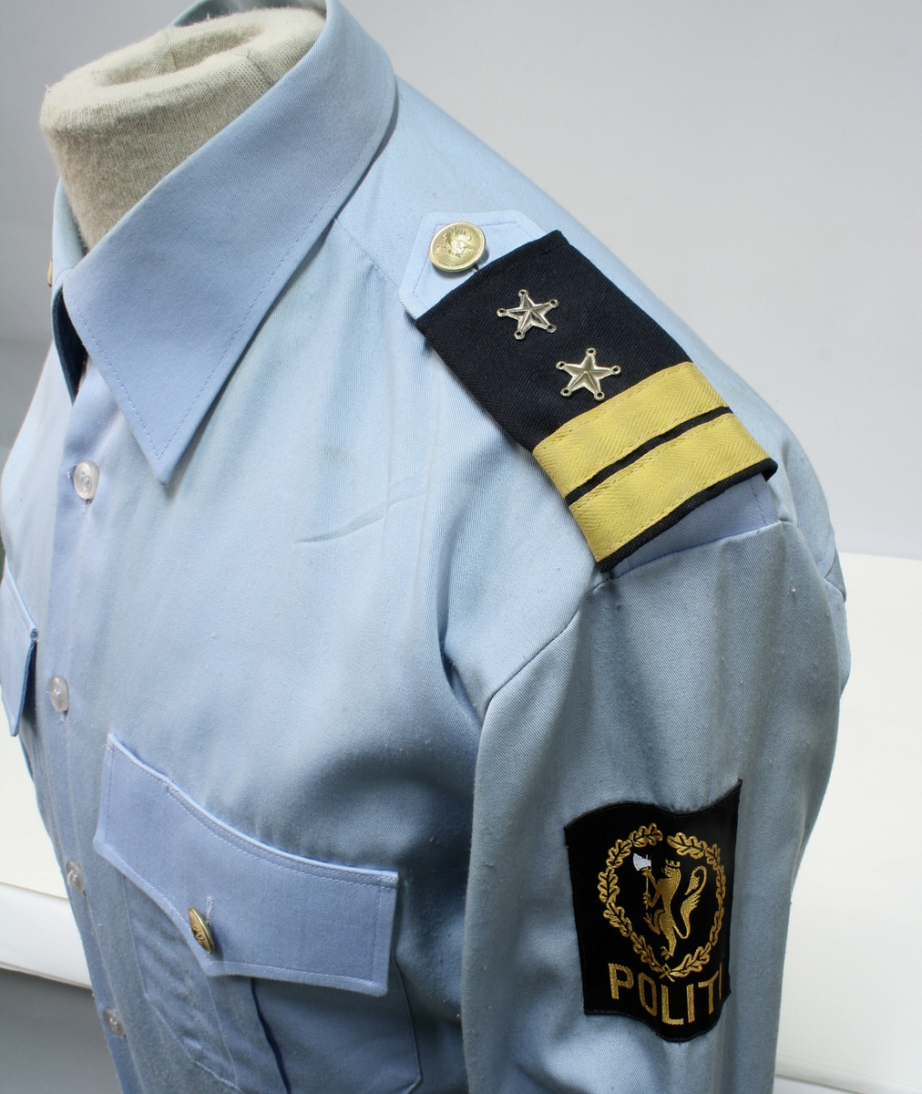 Uniform for politinspektør etter 1963-reglement med merking etter 1977-reglement. Helårsuniform M/1963 med skjorte, langbukse, belte, helårskappe og jakke. Skyggelue med gullbrodert bånd, loslue og båtlue M/1967. Kappen og jakken har skulderklaffer av flettede gullsnorer med to sølvstjerner og festeknapp i gullbroderi. Skjorten har en enklere skulderklaff med to gule render og to sølvstjerner. Det følger med et ekstra enkelt lærbelte og et ekstra sett skulderklaffer til skjorte. Plaggene har gullfargede løveknapper.