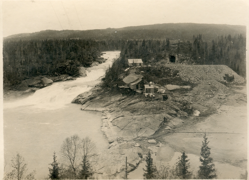 Landskap. Oversiktsbilde. Ei stri elv med foss. Noen bygninger ved fossen, skogkledt landskap. Opplysn; Reinfossen utemmet 1921.