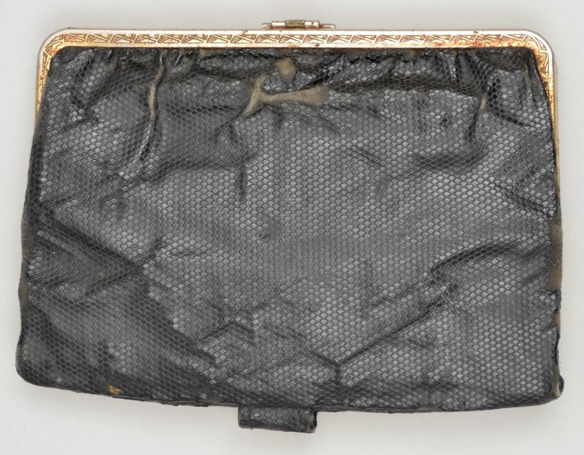 Lita handveske utan handtak. Svart skinnimitasjon. Lukking med metall-lås. Kant i metall. Lomme med knepping inni. Fôra med bomullsstoff. To kalenderlappar med dato 10 og 13 mai 1944 oppi lomma.