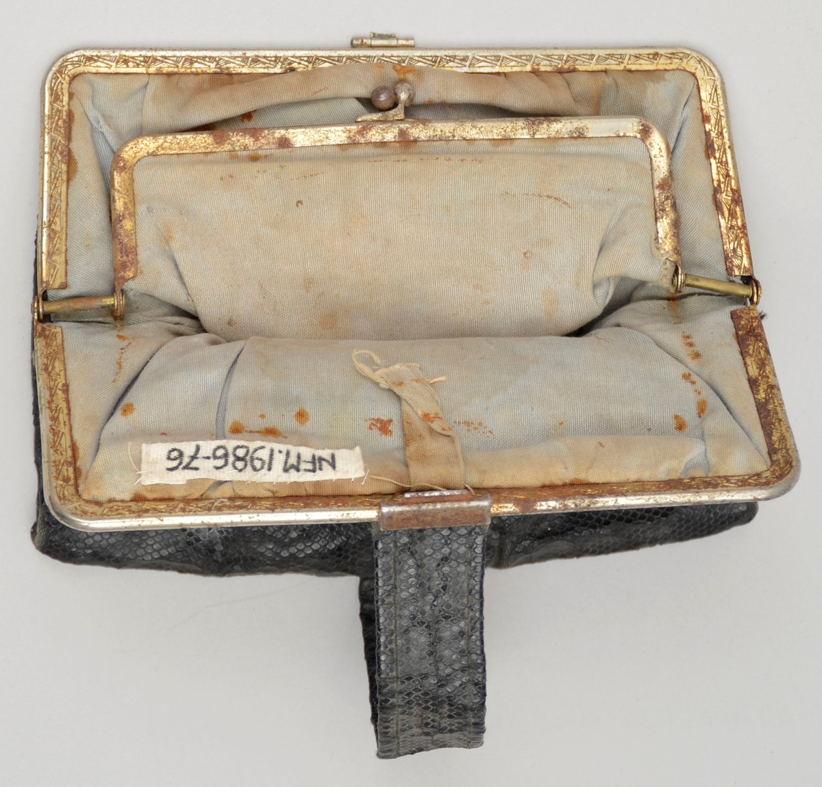 Lita handveske utan handtak. Svart skinnimitasjon. Lukking med metall-lås. Kant i metall. Lomme med knepping inni. Fôra med bomullsstoff. To kalenderlappar med dato 10 og 13 mai 1944 oppi lomma.