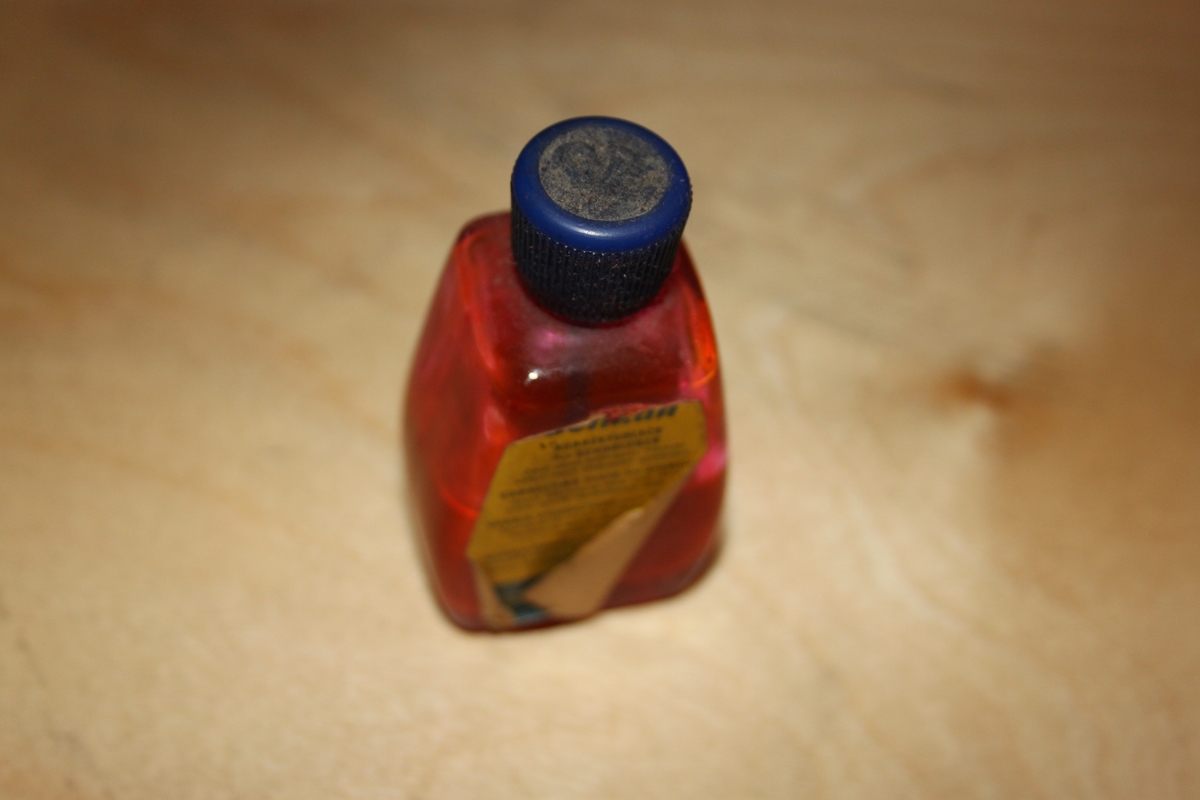 Blekkflaske med skrukork. Glassflaske med papirmerkelapp. Flasken har avrundede former/hjørner. I skrulokket er det festet en pensel.