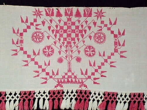 Broderat hängkläde i hallandssöm med botten i tuskaftat vitt linne. Ljusrött (rosa) broderi med tre partier och mönster. Två isydda garnhankar av lintråd i överkanten samt två små näst som inhållning. Påsydd frans i partivis röd och vit bomull med omknutna tofsar längs nederkanten. Frans +:70  mm.