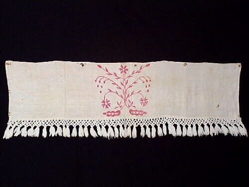 Hängkläde, del av. En liten bit av ett broderat hängkläde med hallandssöm. Bottentyg i tuskaftat vitt linne med ljusrött (rosa) broderi. Påsydd vit frans av bomull med omknutna tofsar i nederkanten. Längd= 100 cm. Bredd= 23,5 cm. Längd (frans)= 8,5 cm.