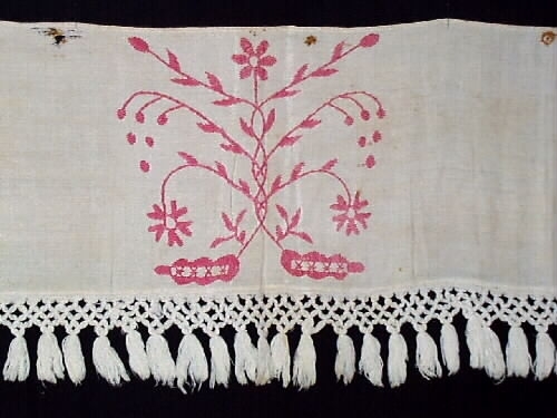 Hängkläde, del av. En liten bit av ett broderat hängkläde med hallandssöm. Bottentyg i tuskaftat vitt linne med ljusrött (rosa) broderi. Påsydd vit frans av bomull med omknutna tofsar i nederkanten. Längd= 100 cm. Bredd= 23,5 cm. Längd (frans)= 8,5 cm.
