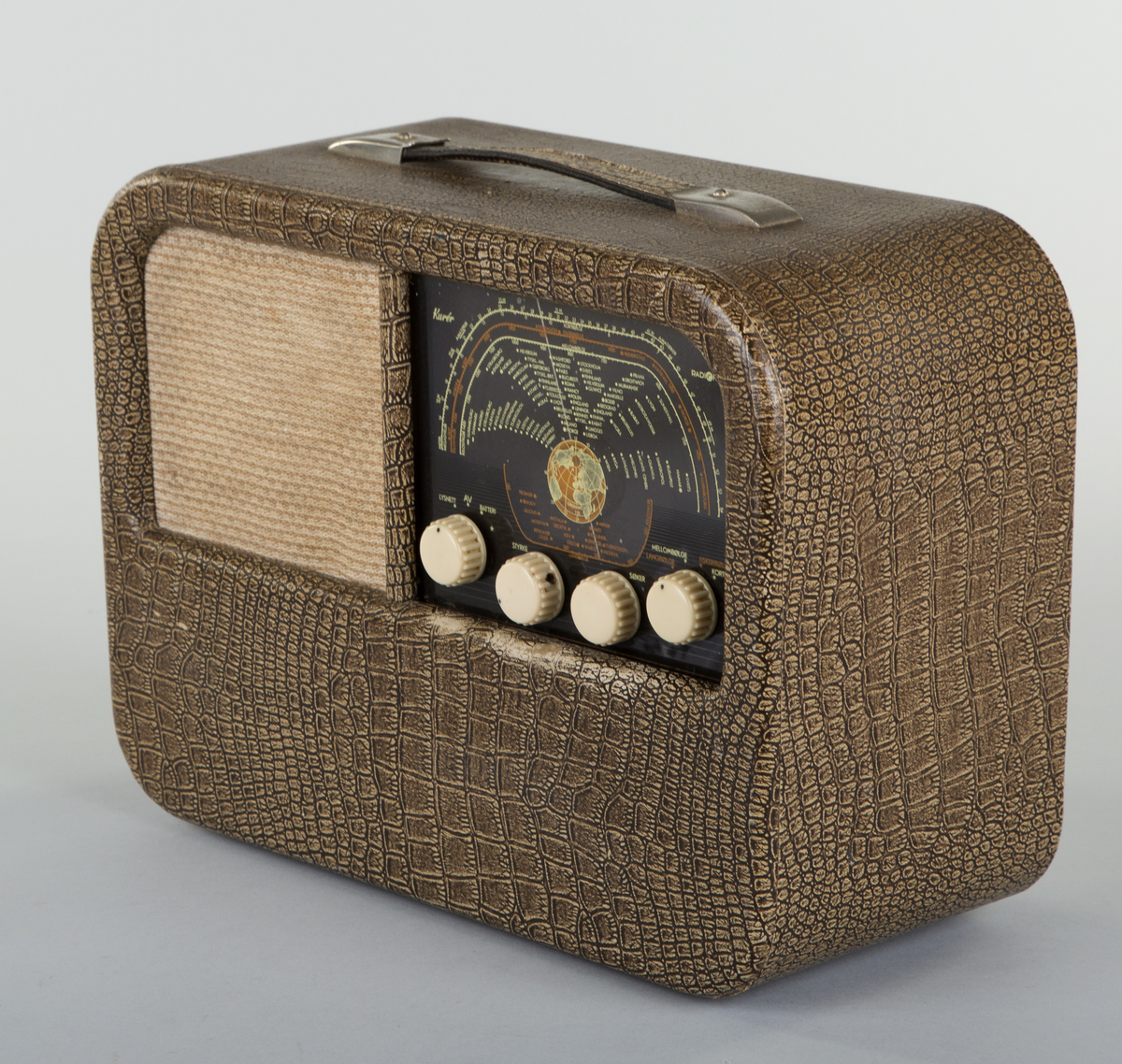 Rektangulær krokodillemønstret reiseradio med runde hjørner. Forside: skala med fire runde funksjons-, og styringsknapper. Høyttaler med trekk av tekstil. Håndtak på oversiden av radioen. Bakside: mangler bakplate. 6 uttak. I tillegg uttak for strøm. Bananstikk til antenne- eller platespilleruttaket ligger vedlagt, innpakket i plast.