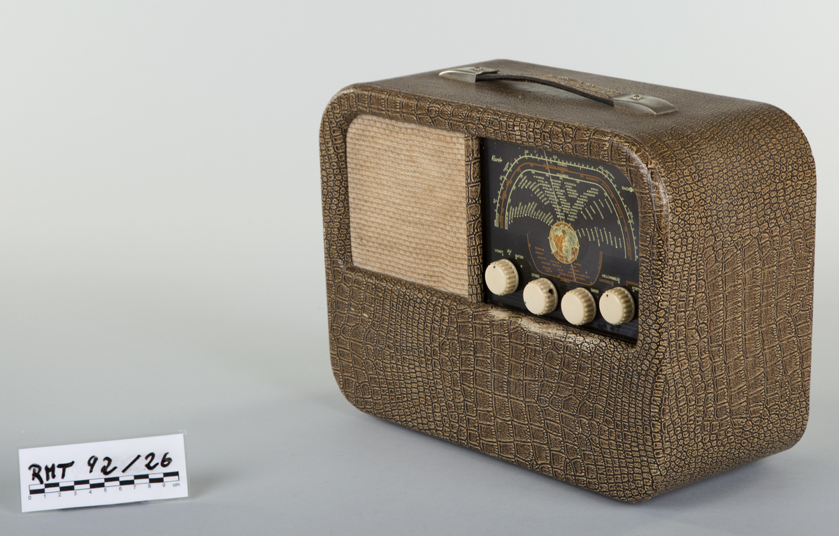 Rektangulær krokodillemønstret reiseradio med runde hjørner. Forside: skala med fire runde funksjons-, og styringsknapper. Høyttaler med trekk av tekstil. Håndtak på oversiden av radioen. Bakside: mangler bakplate. 6 uttak. I tillegg uttak for strøm. Bananstikk til antenne- eller platespilleruttaket ligger vedlagt, innpakket i plast.