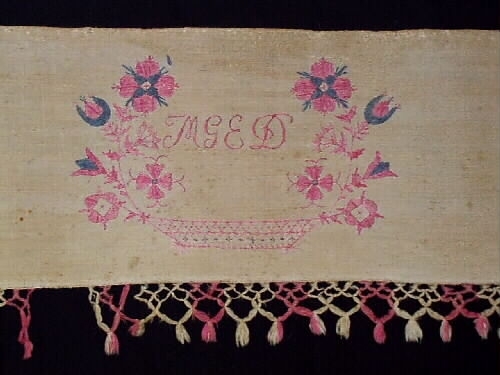 Hängkläde av bomull och linne. Knuten frans i rosa och vitt. Broderier i rosa och blått bomullsgarn. Märkt: M G E D ÅR, 1845.