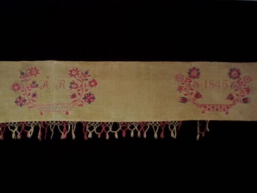 Hängkläde av bomull och linne. Knuten frans i rosa och vitt. Broderier i rosa och blått bomullsgarn. Märkt: M G E D ÅR, 1845.