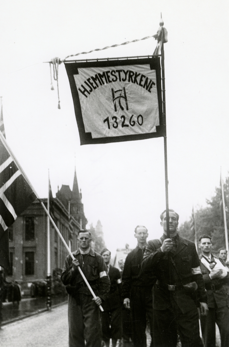 Fra fredsdagene i 1945. Fra Borgertoget.
Hjemmestyrkene marsjerer på Karl Johans gate. Fane med kongens monogram og teksten HJEMMESTYRKENE og nummeret 13260.