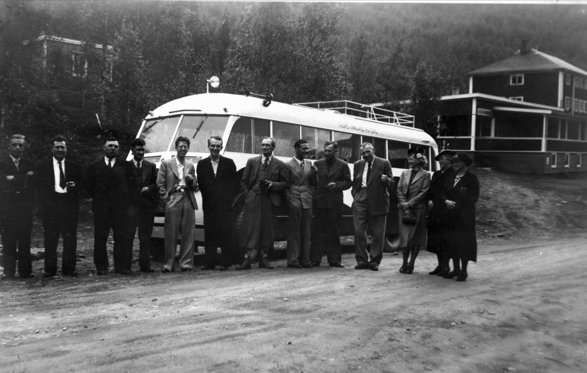 Menn og kvinner foran en buss. Skog og bygninger i bakgrunnen.