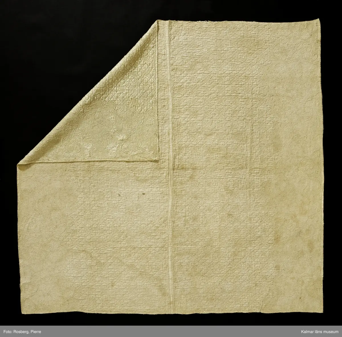 KLM 17625. Täcke av vitt siden med broderi och sticksöm i vitt silke. Utmed två kanter breda bårder med broderade snirkelornament av fransk liljekaraktär. I övrigt rutor med blommor och fyrauddiga snirklade stjärnor. Vitt linnefoder. Tunn bomullsvadd. Långsidorna avklippta. Täcket har varit kantat runtom med vitt sidenband. Troligen omsytt av annat föremål. Sidenet är mycket skadat.