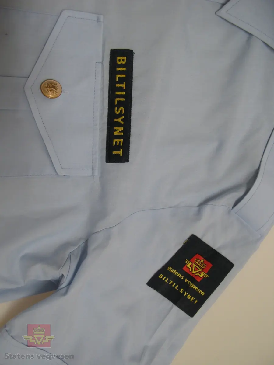 Kortermet lyseblå uniformskjorte i tynt bomullsstoff med skjortekrage, skulderklaffer og enkel knapping. Skjorta er i størrelse 41, herremodell. To brystlommer ,gullfargede knapper (type riksvåpen) både på skulderklaffer og lommer. Tøymerke med "BILTILSYNET" over venstre brystlomme. Påsydd firkantet merke med etatslogo og Biltilsynet, på både høyre og venstre arm.