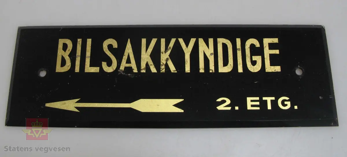 Svart rektangulært skilt med teksten "BILSAKKYNDIGE 2. ETG"
Skiltet har to hull for fastsetting.
