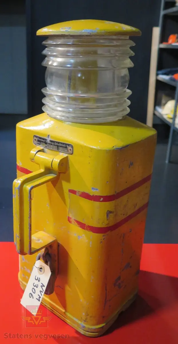Rektangulær varsellykt (lampe). Den er gul med rødes striper. Drevet av fire Hellesens 1,5 volt batterier, seriekoblet til 6 volt, (her fjernet). Tekst "El-fyr".