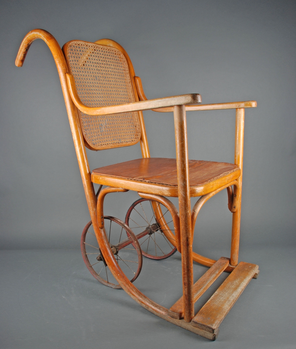 Rullestol i formpresset tre. Fotplate. Håndtak i forlengelsen av ryggen. Rotting rygg. Finert sete erstatter rotting. 2 hjul.