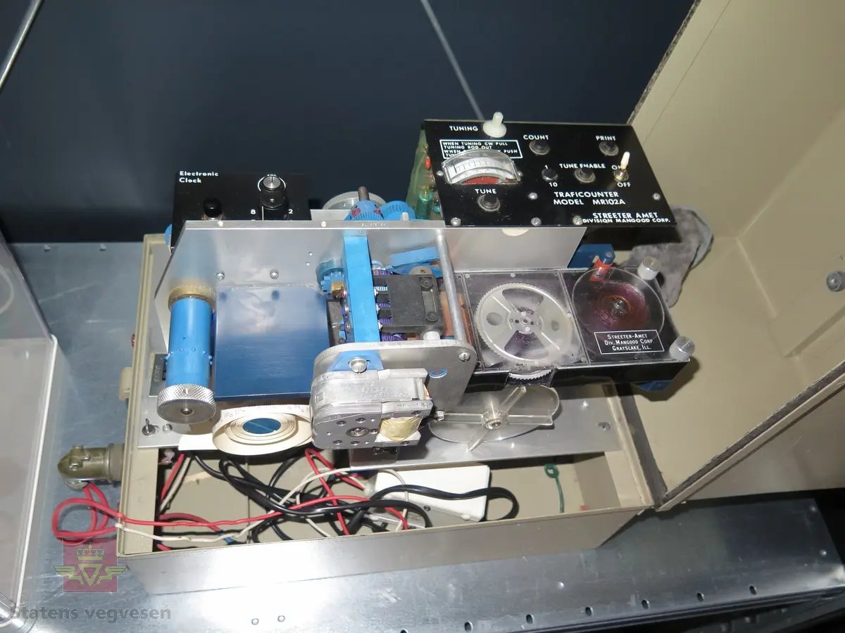 Telleapparat i kasse. Mangler føler. Trafikktelleren drives av 2 stk. 6 volt batterier, av typen Power Sonic Model PS 682, de er 8 ampere og er seriekoblet. Batteriene er nå utkoblet og ligger i kassa. En batterilader med 220 volt kontakt er tilkoblet telleren.