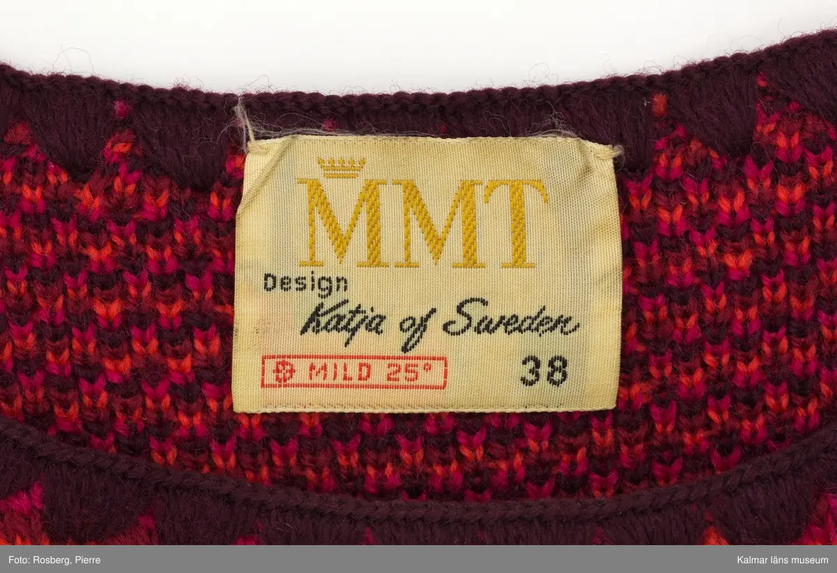 KLM 39302:13. Klänning. 1970-tal. Av jaquardstickad ylle, fyra färgs jaquard med köperbaksida. Mönster; växtdelar i stora rombformer i röda nyanser på orange botten. Knälång, rak modell. Rund halsringning, maskinlaskad med mörkrött garn. Ärmslös. Ärmhål och fåll sydda med overlocksöm. Nära fållen på avigsidan finns en fastsydd remsa av rött fodertyg, troligen rest från ett bortklippt helfoder. Fastsydd textileikett mitt bak med invävd text: MMT Design Katja of Sweden MILD 25 38.