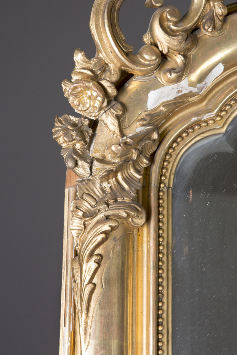 Stort, rektangulært speil med utskjæringer på rammen. Forgylt ramme. Rocailler i asymmetrisk mønster. Speilet buer seg lett ved toppstykket. Rikt asymmetrisk utskjært toppstykke med blomster og rocailler.