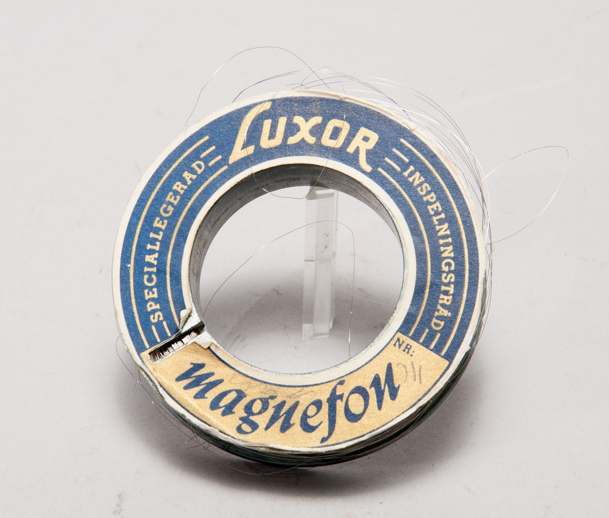 Tontråd till trådspelare, på svarvad metallspole utan etikett, märkt M 22702/60. I pappask med texten: "Luxor Magnefon, talande tråden". På medföljande pappersremsa anges: "Per Lindfors program". Spole: ytterdiameter: 70, håldiameter: 40, tjocklek: 17 mm. Kartongens längd: 80 mm, bredd: 80 mm, höjd: 20 mm.