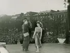 Modell i kjol, linne och ballerinaskor fotograferas av man på en scen i Kungsträdgården, Stockholm.