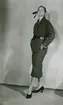 Modell i dräkt med skärp, hatt med dekorband och pumps, av Jacques Griffe.