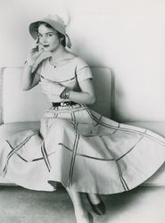 Modell i klänning, hatt, handskar och armband i en soffa. "I