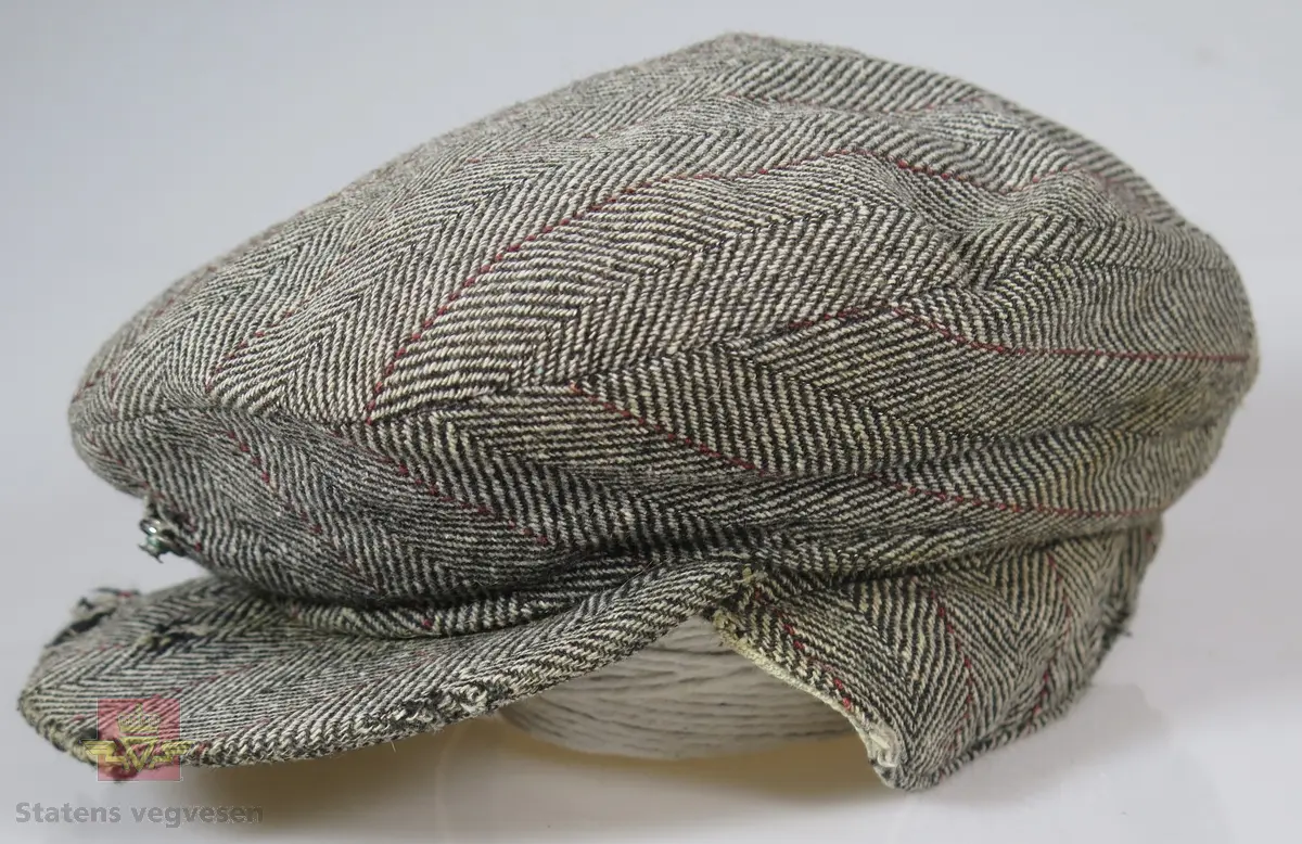 Lua er laget i ullstoff, tweedstoff i fiskebeinsmønster. Hovedfargene er sauebrunt og ubleika med en smal stripe i rødt/rustrød. Skyggen/skjermen er stivet opp med en pappplate. Trykk-knapp holder lua og skyggen sammen. Øreklaffer kan trekkes ned. Vatert silkefôr eggeskallshvit farge. Bl.a. er skyggen noe defekt, dvs med hull. Papplaten i skyggen er delt i to. Lua er vasket etter innkomst til ausjonarius. Lua er ganske slitt.