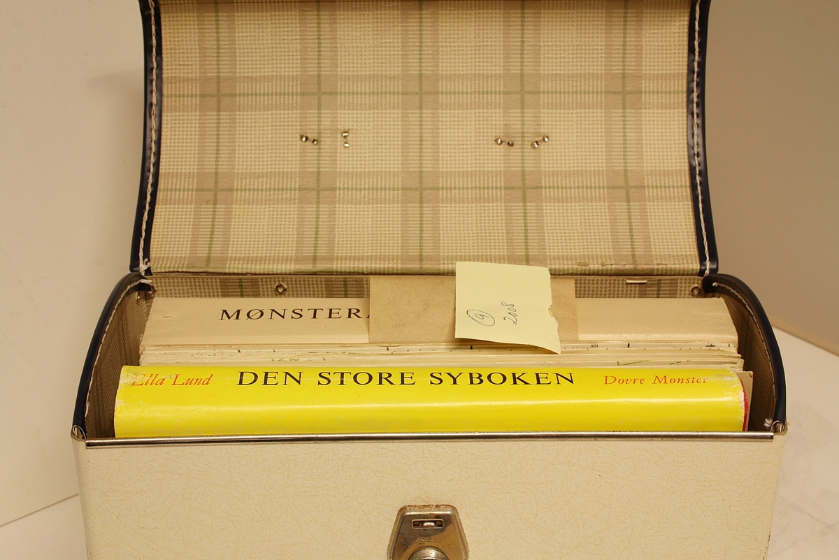 Rektangulær, stående koffert i hvitt og svart med lokk, håndtak på toppen og lås foran. Den inneholder boka "Den Store Syboken" av Ella Lund, samt to mapper med 21 mønsterark i hver med mønster for sying av klær til barn (blå), tenåringer (orange), voksne kvinner (grønn), herrer (svart) og dukker (rød).