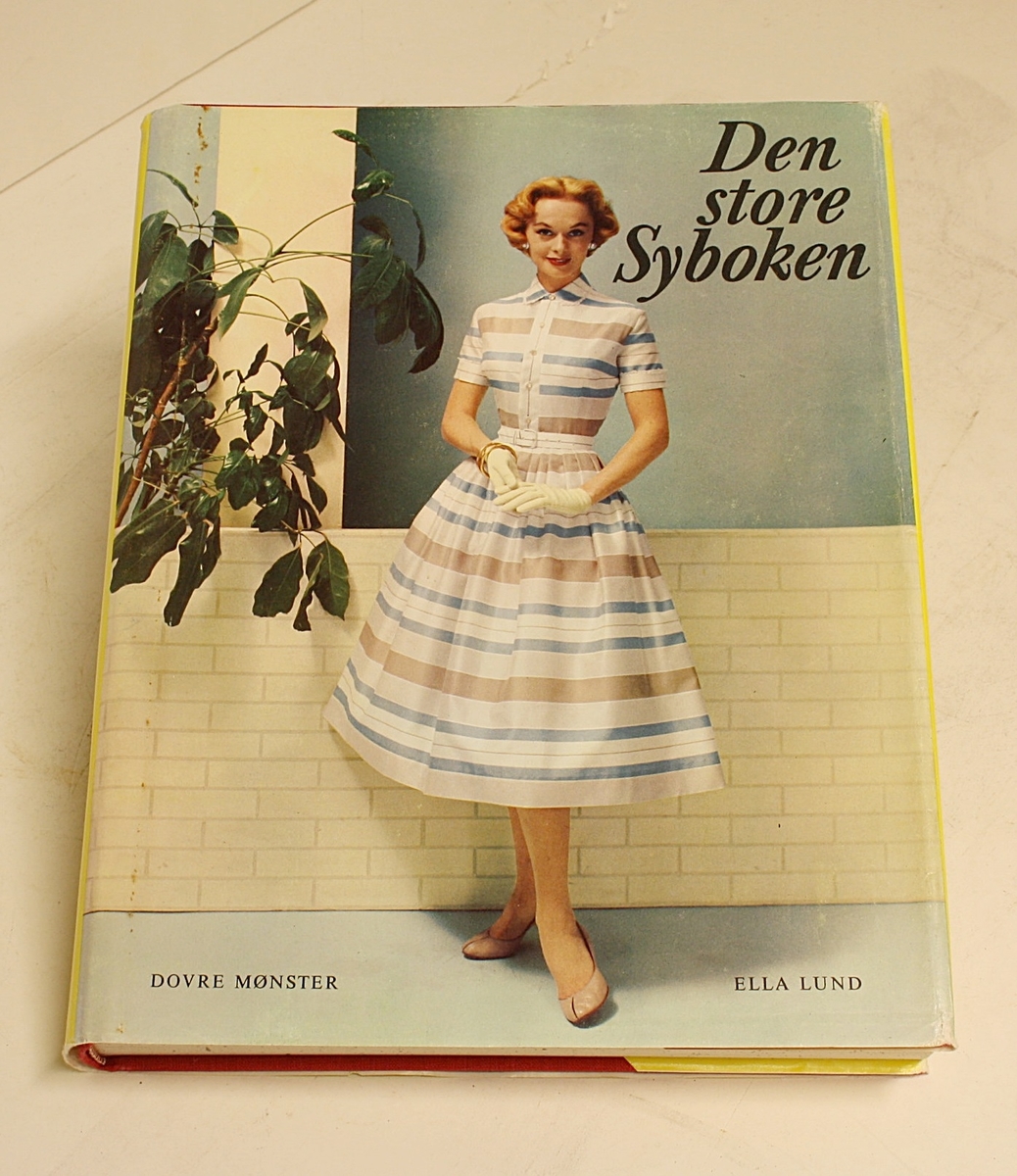 Rektangulær, stående koffert i hvitt og svart med lokk, håndtak på toppen og lås foran. Den inneholder boka "Den Store Syboken" av Ella Lund, samt to mapper med 21 mønsterark i hver med mønster for sying av klær til barn (blå), tenåringer (orange), voksne kvinner (grønn), herrer (svart) og dukker (rød).