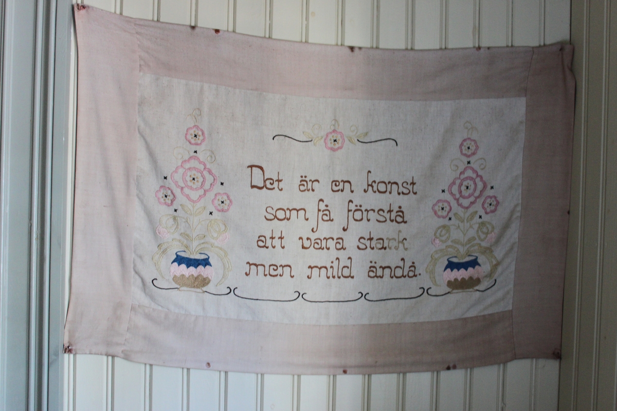 Väggbonad i tyg med broderat motiv och text. På en naturfärgad botten är broderat två blömkrukor med stiliserade blommor omgivande en text. Broderi med stjälkstygn, korsstygn, plattsöm och langettstygn i
färgerna blått, brunt, grönt och rosa