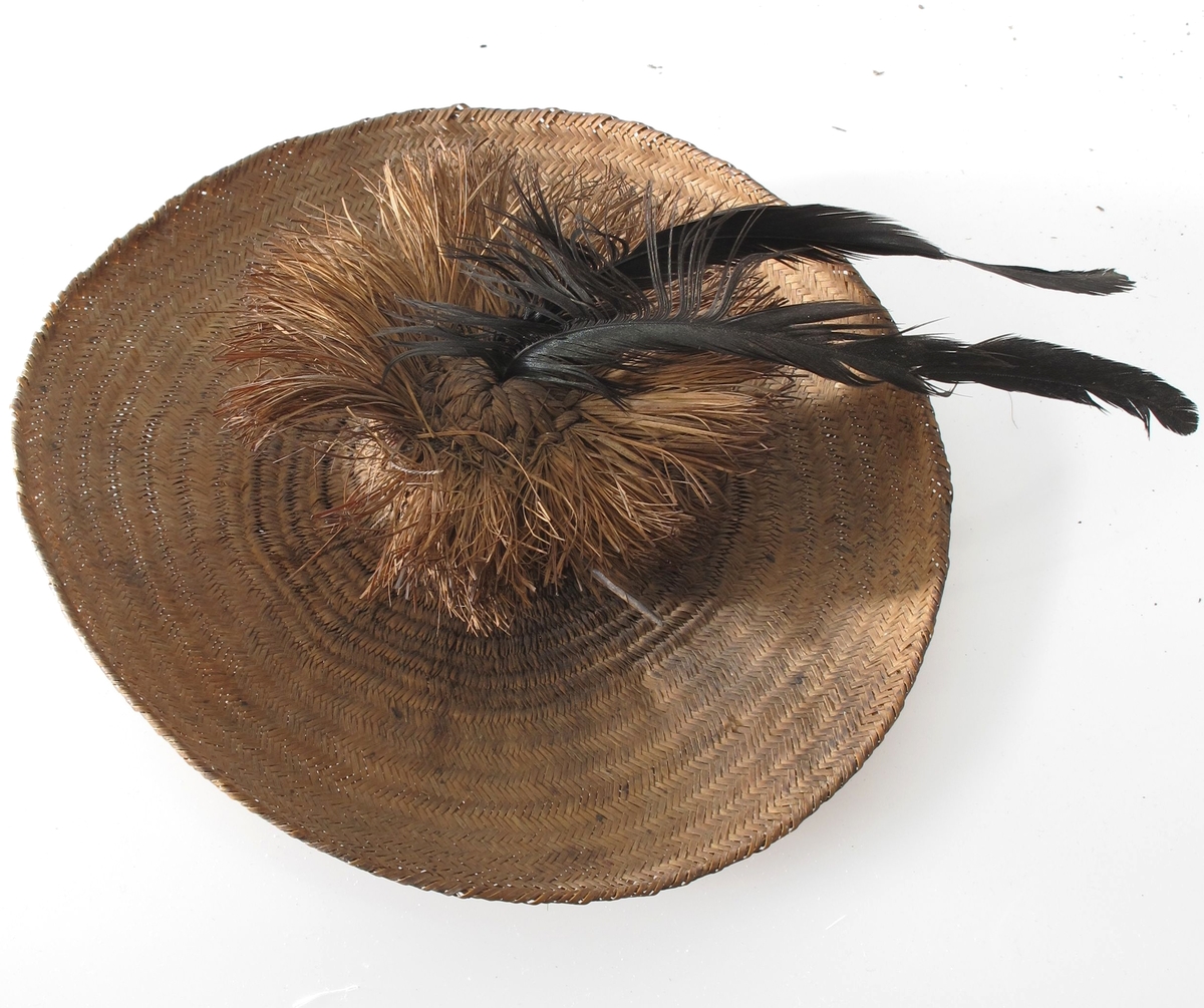 Vidbremmet hatt i mykt strå, brunt, med en liten sylindrisk  pull med  en 4,5 cm lang krans av strå rundt toppen. I toppen en sort  buet  fjær, gjennom pullen en hattenål i form av en fjærstilk.