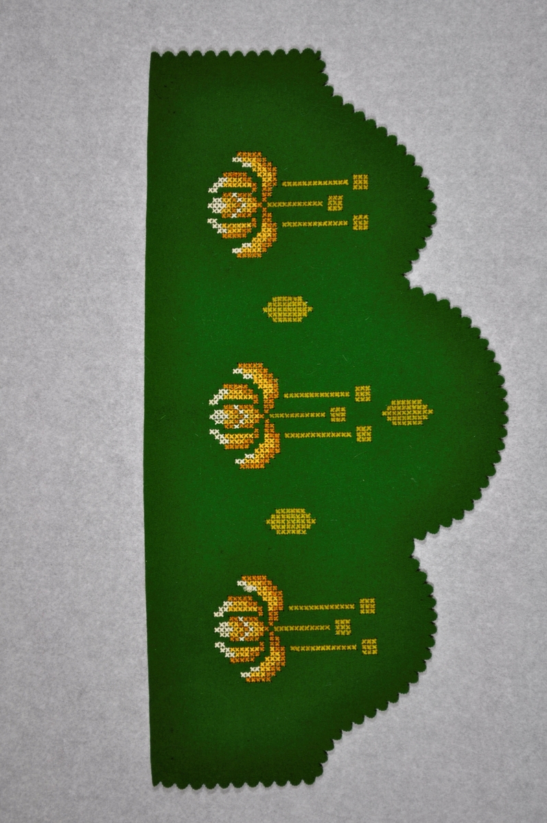 Kappen er laget i grønn filt med korsstingsbroderi i gult og hvitt.
Den er laget som pynt til vegghylle.