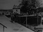 Pråmen ligger i Nykvarns sluss vid Linköping. En av personerna på bilden är pråmskepparen Oskar Broberg från Gottfridsberg. 
Pråmen AGE 4 byggdes 1919 i Linköping och delar av den finns sedan 1953 i strandkanten vid Hjulsbrovarvet.

Övriga anteckningar: Extern information, 2003-12-30, av Joakim Johansson, Linköping (Kinda kanalkännare).