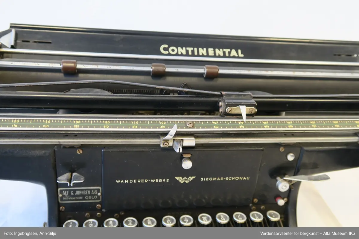 Rektangulær svartlakkert mekanisk skrivemaskin på fire bein med en bred skrivevalse som går langt utover maskinens grunnform. Maskinen har et overtrekk i tekstil.