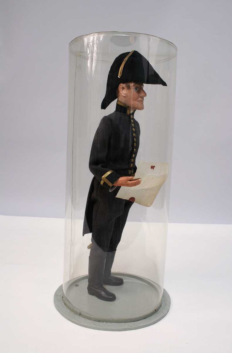 Grå kjole og benklær, grønn krage og oppslag, sorte støvler, sort snutehatt med gullkrampe, sabel med geheng. Montert på sirkelrund plate. Modellen har gradstegn for politiadjutant. Modellen står inni en plastsylinder på en treplate.