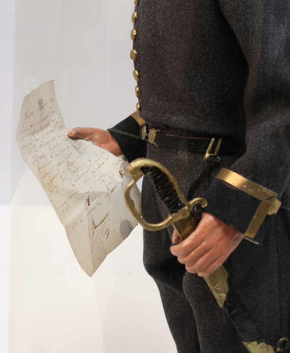 Grå kjole og benklær, grønn krage og oppslag, sorte støvler, sort snutehatt med gullkrampe, sabel med geheng. Montert på sirkelrund plate. Modellen har gradstegn for politiadjutant. Modellen står inni en plastsylinder på en treplate.