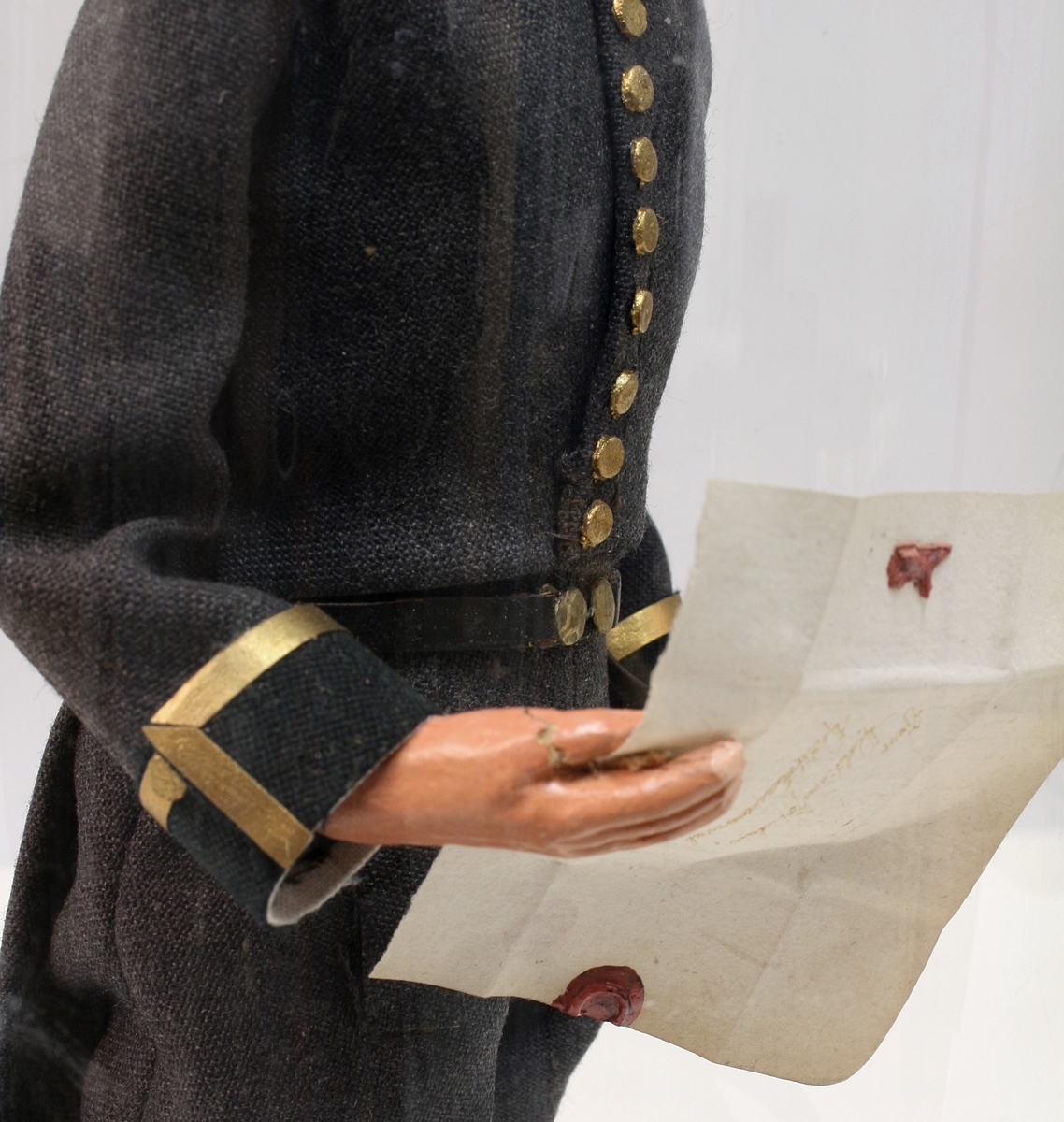 Grå kjole og benklær, grønn krage og oppslag, sorte støvler, sort snutehatt med gullkrampe, sabel med geheng. Montert på sirkelrund plate. Modellen har gradstegn for politiadjutant. Modellen står inni en plastsylinder på en treplate.