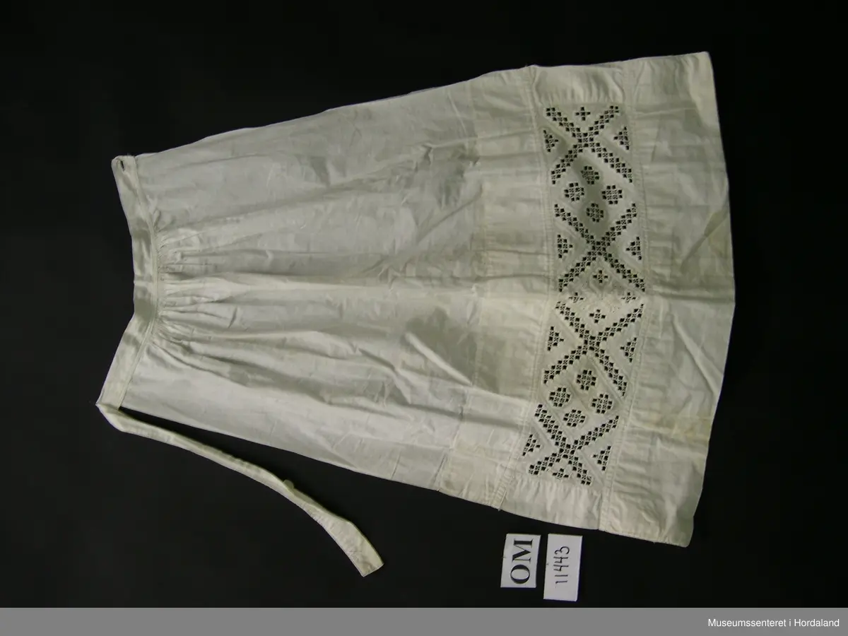 Kvitt bunadsforklede med hardangersaumbord felt inn 14 cm frå nedre kanten. Motivet er inndelt i firkantar, i midten framme er det sydd ei åttebladrose. Linning i livet som fortset rundt ryggen og knappast i sida. Forkledet er rynka ved linninga midt framme. Ikkje rynka i sidene.