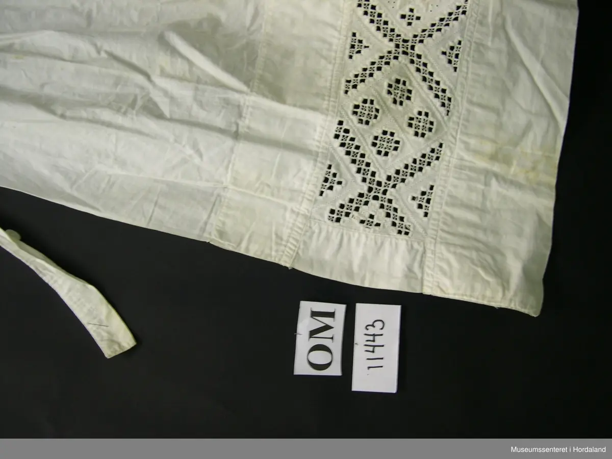 Kvitt bunadsforklede med hardangersaumbord felt inn 14 cm frå nedre kanten. Motivet er inndelt i firkantar, i midten framme er det sydd ei åttebladrose. Linning i livet som fortset rundt ryggen og knappast i sida. Forkledet er rynka ved linninga midt framme. Ikkje rynka i sidene.