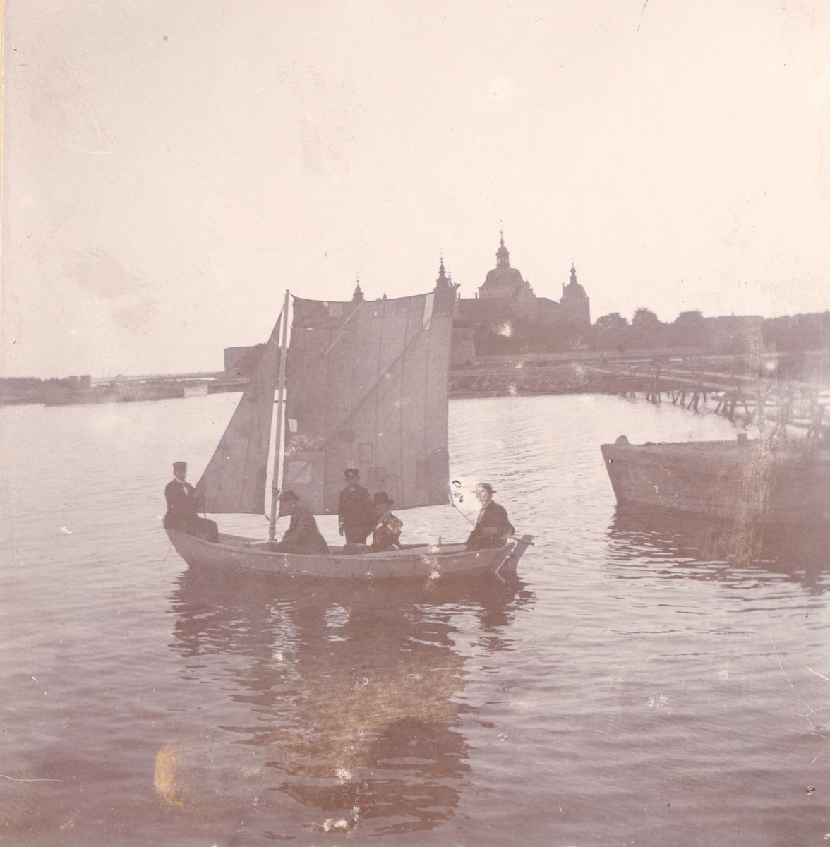 Flera män sitter i en segelbåt i slottsfjärden med Kalmar slott i bakgrunden. Bilden är troligen tagen runt sekelskiftet 1900.