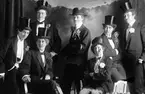 Fröken Dahlgren och fröken Forsberg mfl i kostymering. Dahlgren är antingen kvinnan på bakre raden längst till höger eller kvinnan som sitter i stolen till vänster, detsamma gäller för Forsberg.