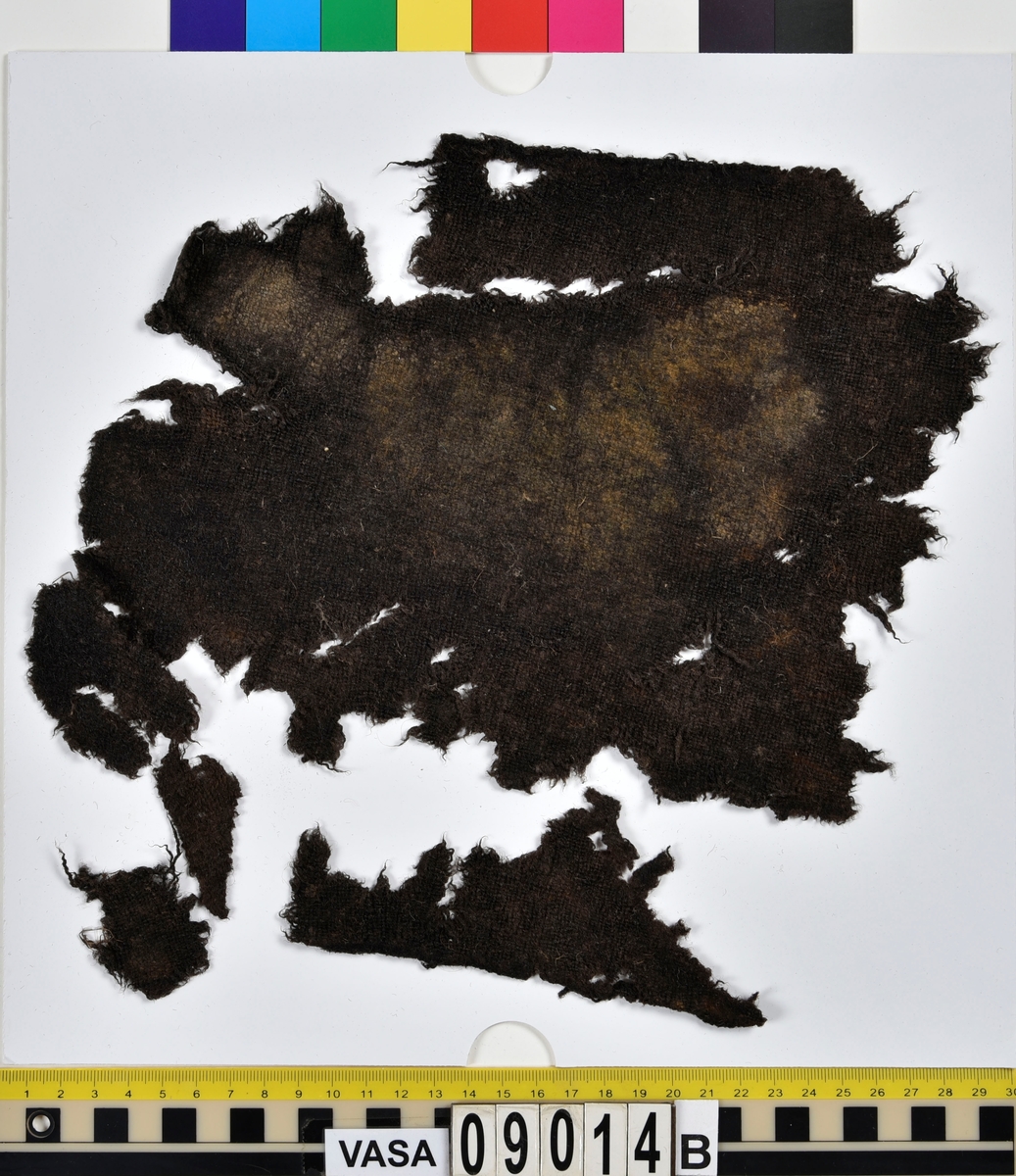 Textildokumentation 2018
Textil, fyndnumret består av mer än 50 st fragment uppdelade på 09014a-h.
Kontextgrupp ÖB20 (övriga fyndnummer i gruppen: 07732, 07762, 09228)
Relaterade fynd: Kontextgrupp ÖB17 och ÖB 18.
.
Fnr 09014a Typnr ÖB20.3:1
Sammanfattning - Ett textilfragment av ull vävt i tuskaft. Färgen är nu mörkbrun, fibrerna varierar i färg vilket tyder på att det är naturfärgad ull. Fibrerna är i vissa partier skadade på ytan och ser därför vita ut. Väven är tunn, jämn och fin och har inga spår av ytbehandling. Fragmentet har tre tillskurna kanter, två av dessa har spår av tre olika sömmar (söm 1: kvicksöm/förstygn). Formen tyder på att det är ett skört till en tröja. Andra delar: Jämför med 07726. 
.
Fnr 09014a Typnr ÖB20.4:1
Sammanfattning - Ett textilfragment av ull vävt i tuskaft. Färgen är nu mörkbrun, originalfärg okänd. I mikroskop ser tyget blåsvart ut. Väven är mycket jämn och slät. I sömsmånen på sida A finns bevarad lugg, på sida B är luggen så välbevarad att bindningen inte syns. Fragmentet har två tillskurna kanter med hål och avtryck av sömmar, även trådrester finns (söm 1: kaststygn - fälld kant, söm 2: ev förstygn - sammanfogande, söm 3: kaststygn - fälld kant). På sida B finns blåfärgad ull och silkesfibrer. 
.
Fnr 09014a Typnr ÖB20.5:1
Sammanfattning - Ett textilfragment av ull vävt i tuskaft. Väven är jämn och tät. En av kanterna är troligen tillskuren, inga sömrester finns. Tyget har inga spår av ytbehandling. På ytan finns en del bastfibrer. Relaterade fynd: Vävens struktur liknar ÖB20.4:1 men tygerna har ändå olika karaktär och detta fragment saknar lugg. 
.
Fnr 09014a Typnr ÖB20.6:1
Sammanfattning - Ett textilfragment av ull vävt i tuskaft. Färgen är nu brun, originalfärg är okänd men möjligen naturfärgad, fibrerna är mycket släta och varierar i färg från brun till nästan svart. Fragmentet är litet och mycket skadat, endast några få bindepunkter återstår vilket försvårar mätningar. 
.
Fnr 09014a Typnr ÖB20.7:1
Sammanfattning - Ett textilfragment av  ull vävt i tuskaft. Färgen är svartbrun, originalfärg okänd men möjligen naturfärgad då fibrernas färg varierar. Inga tydliga spår av ytbehandling. Väven är jämn men garnet är hårt spunnet vilket ger tyget en kypertkaraktär (falsk kypert). Fragmentet har inga tillskurna kanter eller sömmar. På ytan finns enstaka vita silkesfibrer.
.
Fnr 09014a Typnr ÖB20.8:1
Sammanfattning - Ett textilfragment av ull. Fragmentet är mycket litet och dåligt bevarat så bindningen är svår att avgöra, troligen 2/2 kypert. Färgen är svart, möjligen naturfärgad ull då fibrerna varierar något i färg. Tyget har ingen synlig ytbehandling. Garnet är hårt spunnet vilket ger tyget en speciell karaktär. 
.
Fnr 09014b Typnr ÖB17.1:8-11
Sammanfattning - Fyra textilfragment av ull vävt i tuskaft. Färgen är mörkbrun, originalfärg okänd men den bruna tonen slår mot röd. Tre av fragmenten har tillskurna kanter och spår av sömmar. Andra delar, som troligen kommer från samma tröja, är fnr 03261a och d, 09014h samt 09228a. 
Fragment ÖB17.1:9 - Fragmentet har två tillskurna kanter varav den ena har spår av en söm. Fragmentet är troligen sidostycket till en tröja. Den tillskurna kanten har då varit monterad mot tröjans bakstycke och bör ha hört samman med fyndnummer 03261d, vilket är ett bakstycke till en tröja.
Fragment ÖB17.1:10 - Fragmentet är mycket skadat men är troligen av typen ÖB17.1. Formen är densamma som för del av fragment ÖB17.1:9 och kan därför vara en dubblering av detsamma, alternativt det motstående sidostycket.
.
Fnr 09014c Typnr ÖB20.9:1-19
Sammanfattning - Nitton textilfragment av ull vävt i tuskaft. Färgen är brun, originalfärg okänd. Sida B har en kort tät lugg som döljer bindningen. På sida A finns ingen lugg och bindningen är synlig. Fjorton av fragmenten har tillskurna kanter och sömmar. Tre av fragmenten har sydda knapphål. Flera fragment är dräktdelar, till exempel finns en fragmentarisk krage. På ytan på flertalet fragment finns fibrer i flera klara färger, till exempel rosa, röd och blå. Vita fibrer finns i stor mängd, kan vara recenta.

Fragment ÖB20.9:1 - Fragmentet har flera tillskurna kanter med avtryck och hål efter olika typer av sömmar (söm 1: ev. sammanfogande, söm 2: kaststygn - fåll, söm 3: kaststygn - sammanfogande, söm 4: kaststygn - sammanfogande, söm 5: osäkert, söm 6: osäkert), trådrester finns. Kanterna och sömmarnas karaktär gör att sida A bedöms vara plaggets rätsida/utsida. Några hål som ej är stygnhål men kan vara original finns. I fragmentet sitter fast två stycken silkestrådar (söm 7: silkestråd med knut, fäst i tyget, söm 8:  silkestråd med ögla, fäst i tyget). Det är samma typ av tråd som knapphålen på fragment ÖB20.9:2 är sydda med. På sida A finns ett område med extra mycket korrosion och även en långsmal vitaktig avlagring.

Fragment ÖB20.9:2 - Fragmentet har inga bevarade tillskurna kanter, dock finns en någorlunda välbevarad rad med knapphål. Samtliga knapphål är trasiga i ena sidan så deras ursprungliga storlek går ej att avgöra. Knapphålen är sydda med knapphålsstygn i silkestråd och sitter med 13-15 mm mellanrum (c/c mått). Det tvärställda tränsen består av ca 10 stygn och syns ej på sida B, tränsens knutar sitter vända från knapphålet. På sida B anas rester av bastfiber under knapphålsstygnen, troligen från en dubblering av tyget eller ett foder. 

Fragment ÖB20.9:3 - Fragmentet har fyra stycken knapphål, varav ett är helt. Knapphålen är skurna på skrådden. Eventuellt finns spår av en söm längs en del av kanten. Knapphålsstygn och trådrester är mycket fragmentariska.

Fragment ÖB20.9:4 - Fragmentet har tre stycken knapphål.  Knapphålen är sydda med knapphålsstygn i silkestråd. Knapphålens längd är 19-20 mm och de sitter med 18-22 mm mellanrum (c/c mått). Eventuellt är fragmentets ena kant en sömsmån, kan ha varit monterad mellan andra tyglager (kanten har avvikande färg). Under knapphålsstygnen på sida B finns rester av annat material, eventuellt bastfiber. Bastfibrer finns också utspridda över ytan på sida B, kan vara rester av en dubblering till tyget eller ett foder, se fragment 9:2. På sida A finns avlagringar och korrosion. 

Fragment ÖB20.9:5 - Fragmentet har bristningar i tyget som ser ut som knapphål, dock finns inga rester av stygnhål eller tråd. Fragmentets ena kant är troligen en sömsmån, kan ha varit monterad mellan andra tyglager (kanten har avvikande färg). Enstaka spår av söm och tråd finns längs kanten (söm 1), dessa är mycket otydliga. På fragmentets andra (mycket trasiga) kant finns ett mycket tydligt stygnhål. Fragment 9:4 och 9:5 ser ut att kunna höra ihop, dock går trådsystemen åt olika håll så det är inte fallet.

Fragment 9:6-9 - Till det stora fragmentet 9:6 hör det lilla lösa fragmentet 9:7. I fragment 9:6 sitter två fragment fastsydda, 9:8 och 9:9. De sammansydda fragmenten har tre tillskurna kanter och spår av minst fem sömmar (söm 1: kaststygn, söm 2: förstygn el. liknande - sammanfogande, söm 3: kaststygn, söm 4: sammanfogande, fäster ihop 9:6 med 9:8 och 9:9, söm 5: två stygnhål), trådrester finns. Det är troligt att 9:8 och 9:9 är delar av samma tillskurna bit, och att den utgjort en dubblering till 9:6 (inklusive 9:7). Den tillskurna formen ser ut att vara en krage. Fragmentet skulle då vara en halv krage, bevarad från framkant till nästan mitt bak. På sida A finns en beläggning på delar av ytan. På sida B finns en mängd fibrer i klara färger: blå, vit, rosa, tegelröd, orange, karminröd, grön, mörkgul och grå. fibrerna ligger ytligt med nästan samtliga sitter delvis fast i tygets lugg eller har en eller båda ändar nere i vävens bindning. Fibrerna bör därför vara ursprungliga. Sida A har en mindre mängd av nämnda fibrer, mest förekommande på ytan av fragment 9:8 och 9:9.

Fragment ÖB20.9:10 - Fragmentet har två tillskurna kanter. Dessa är invikta mot sida B som en 6-7 mm bred fåll som är fastsydd med kaststygn, trådrester finns (söm 1 och 2). Fragmentet kan vara ett skört till en tröja. På båda sidors yta finns fibrer i klara färger: grå, ljusblå, orange, orangebrun, vitgul, blå svart, vit, röd, korallröd och rosa.

Fragment ÖB20.9:11 - Fragmentet har tre tillskurna kanter med sömmar. Två av kanterna är invikta och fastsydda mot sida B, som fållar (söm1: kaststygn - fåll, söm 2: kaststygn - fåll). Stygnen går ej igenom till sida A men sömmen syns som bucklor i fragmentets yta. Söm 3 längs den tredje kanten har troligen varit en sammanfogande söm sydd med kaststygn. Stygn och trådrester finns bevarade från samtliga sömmar. Under fållen vid söm 1 finns ett litet fragment av en annan textiltyp. Troligen ursprungligen en dubblering eller liknande. Fragmentet är ca 10x5 mm stort och har fått typnummer ÖB20.16:1. På ytan på sida B finns enstaka bastfibrer.

Fragment ÖB20.9:12 - Fragmentet har två tillskurna kanter med sömmar (söm 1: ev kaststygn, söm 2: osäker). Vid söm 2 är sömsmånen vikt mot sida B. Vid söm 1 är kanten inte vikt. Fragmentet har samma form som ÖB20.9:6, "kragen".

Fragment ÖB20.9:13 - Fragmentet har två tillskurna kanter med sömmar (söm1: sammanfogande, söm 2: osäkert). De två övriga kanterna är eventuellt också de tillskurna men spår av eventuella sömmar är för otydliga. Enstaka fibrer med klara färger (rosa, blå, vit) finns på båda sidor, fler på sida B. Vid söm 2 finns på ett ställe en fiberknippa som kommer upp ur vävens bindning, kan vara rester av en tråd och är eventuellt halsplat silke. 

Fragment ÖB20.9:14 - Fragmentet har en tillskuren kant med söm (söm1: ev sammanfogande). I stygnhålen finns rester av tråd.
.
Fnr 09014c Typnr ÖB20.10:1
Sammanfattning - Ett textilfragment av ull vävt i tuskaft. Färgen är brun, originalfärg okänd. Sida B har en kort och tät lugg, bindningen syns bara på sida A. Längs ena kanten finns tre stycken tydliga hål. Hålen är gjorda med ett verktyg som skär av fibrerna snarare än tränger undan dem.
.
Fnr 09014c Typnr ÖB20.16:1
Sammanfattning - Fragmentet är mycket litet och sitter inuti den invikta fållen på fragment 09014c ÖB20.9:11, det sticker bara fram någon millimeter. Fragmentet är mycket skört. Bevarandegrad och storlek gör mätningar svåra. Det är olika garn i trs 1 och trs 2. Sida B är täckt av en lugg, på sida A syns bindningen, om än fragmentariskt.
.
Fnr 09014d Typnr ÖB18.16:2-8
Ej dokumenterad
.
Fnr 09014d Typnr ÖB20.11:1-2
Ej dokumenterad
.
Fnr 09014e Typnr ÖB20.12:1-4
Sammanfattning - Fyra textilfragment av ull vävt i tuskaft. Färgen är brun med inslag av täckhårsfibrer i en mörkare brun färg, originalfärg okänd. Fragmenten är mycket fragmentariska med trs 1 bättre bevarat än trs 2. Enbart på en del av fragment ÖB20.12:1 finns trs 2 samt bindningen bevarad. Utspridda över tygets yta finns grova bastfibrer samt korrosionsfläckar. 
.
Fnr 09014f Typnr ÖB20.13:1
Sammanfattning - Ett textilfragment av ull vävt tuskaft. Färgen är mörkbrun, fibrerna varierar mellan ljus och mörk brun. Originalfärg okänd, möjligen naturfärgad ull. Garnet är hårt spunnet vilket ger väven en särskild karaktär, sida A har en mer strukturerad ytan än sida B. Fragmentet har inga tillskurna kanter. På sida A finns enstaka röda och gulgröna silkesfibrer, samt bastfibrer. På sida B finns enstaka blå, vita och rosa silkesfibrer.
.
Fnr 09014g Typnr ÖB20.14:1-3
Sammanfattning - Tre textilfragment av ull och bastfiber vävt i tuskaft. Färgen är beige för bastfibern (trs 1) och svartblå för ullen (trs 2). Trs 2 är mycker grövre än trs 1. Trs 1 är nästan helt försvunnet. Två av fragmenten har tillskurna kanter och ett har spår av en sömsmån. På ytan av fragmenten finns en stor mängd bastfiber samt en vit/rosa tuss med silke. På fragment 14:2 finns några intensivt blå ullfibrer.
.
Fnr 09014h Typnr ÖB17.1:12-13
Sammanfattning - Två textilfragment av ull vävt i tuskaft. Färgen är brun, originalfärg okänd. Det större fragmentet är tillskuret och har spår av sömmar. Fragmentet är en ensömsärm med sömmen placerad mitt bak på ärmen. På ytan finns stora rostfläckar. Andra delar: Hör troligen (trots färgskillnad) till skelettet Beatas tröja, se fnr 03261a-d, 09228a, 09014b och h.