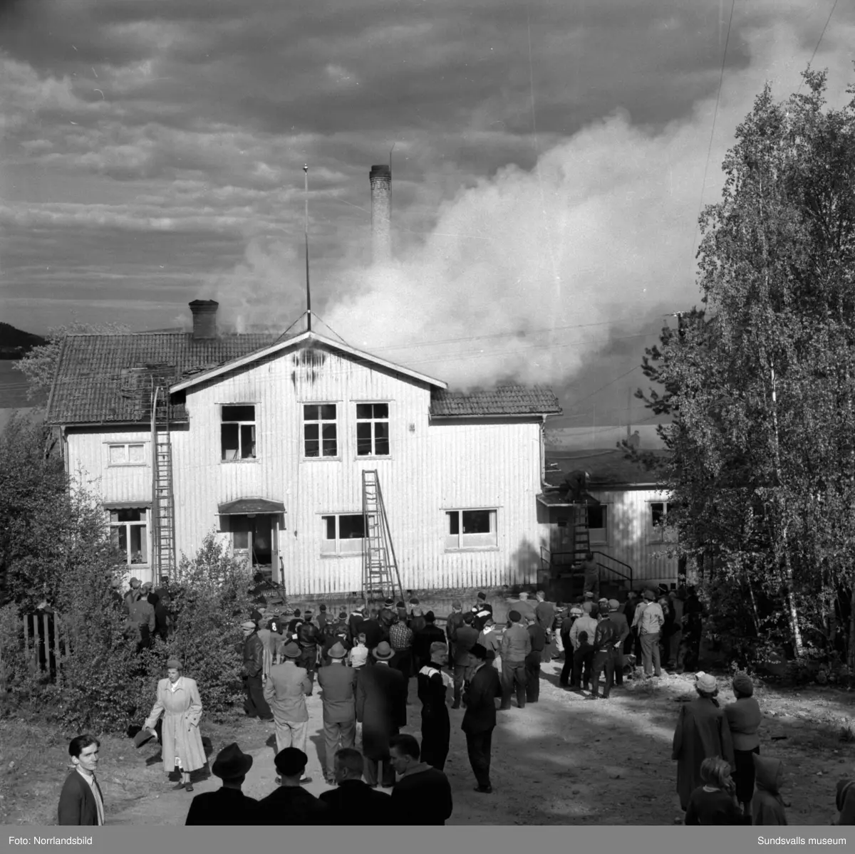 En stor serie bilder från en eldsvåda i kontorsbyggnaden och Bylanders Café vid Luckstaverken, Lucksta ångsåg. Släckningsarbete pågår och massor av människor hjälps åt att rädda så mycket lösöre som möjligt. Elden startade i på övervåningen i kassören Sven Perssons bostadslägenhet. Persson räddade sig ut genom att hoppa från balkongen på baksidan. Brandkårerna från Attmar, Tuna och Sundsvall lyckades till slut få bukt med elden men övervåningen totalskadades och det kafé som fanns inrymt i bottenvåningen fick stora brand- och vattenskador.