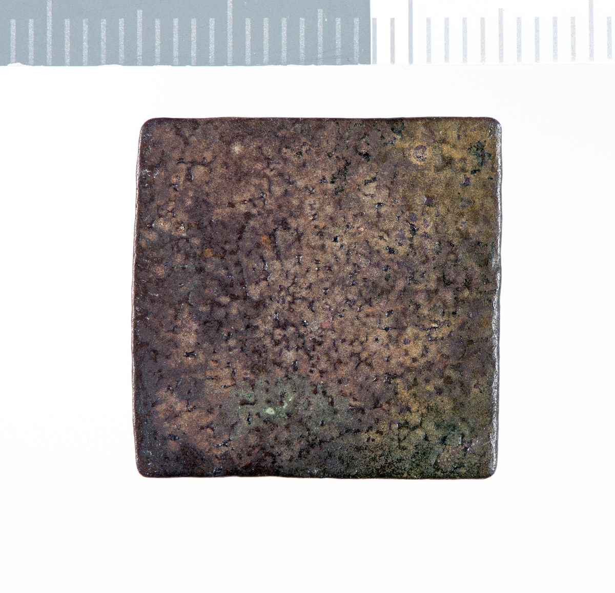 Kvadratisk metallplate med korrodert og  slitt overflate. Sider og hjørner er filt til. Det er rester av et preg på den ene flaten.
