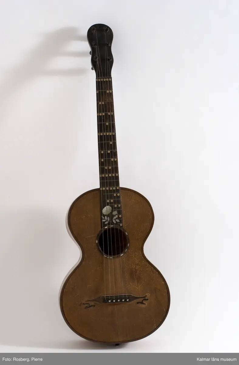 KLM 39707:1. Gitarr, av trä. Inlagd dekor av pärlemor i form av en ros med blad, på greppbrädets nedre del. Komplett med strängar i metall.