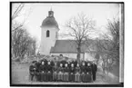 Rinkaby kyrka i bakgrunden.
Konfirmander, 19 ungdomar och en präst.